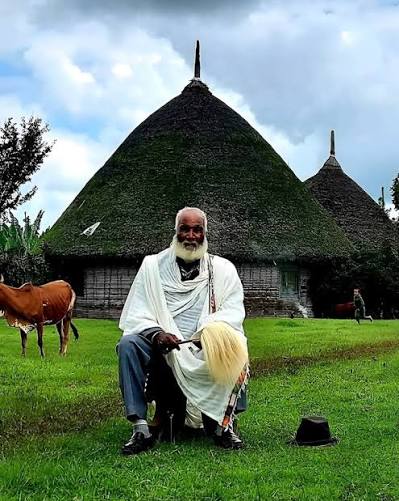 An elder in traditional white dress seated before thatched roundhouses